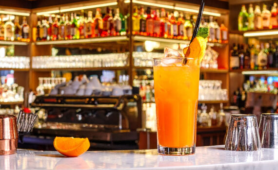 Ein hohes Glas mit einem leuchtend orangen Cocktail steht auf einer weißen Marmortheke. Daneben liegt eine Orangenspalte. Im unscharfen Hintergrund ist eine gut sortierte Bar mit zahlreichen Spirituosenflaschen zu sehen.