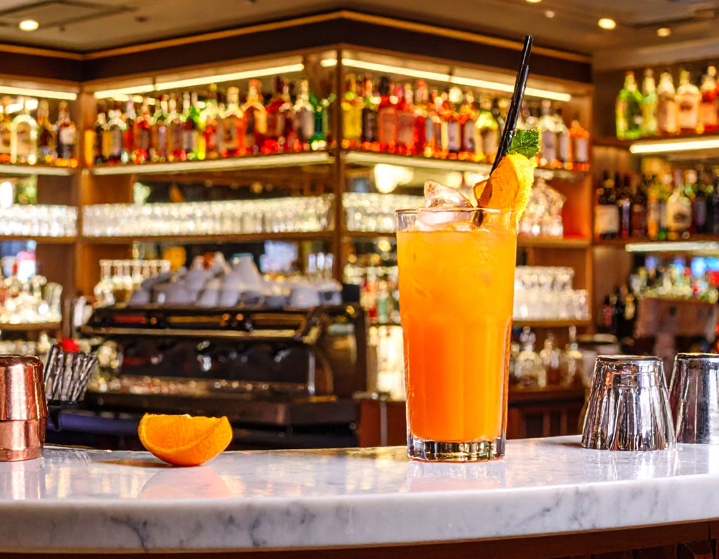 Ein hohes Glas mit einem leuchtend orangen Cocktail steht auf einer weißen Marmortheke. Daneben liegt eine Orangenspalte. Im unscharfen Hintergrund ist eine gut sortierte Bar mit zahlreichen Spirituosenflaschen zu sehen.