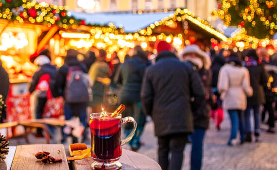 Eine dampfende Tasse Glühwein mit Zimtstange und Sternanis steht im Vordergrund auf einem Holztisch. Im unscharfen Hintergrund erstreckt sich ein belebter Christkindlmarkt bei Abenddämmerung