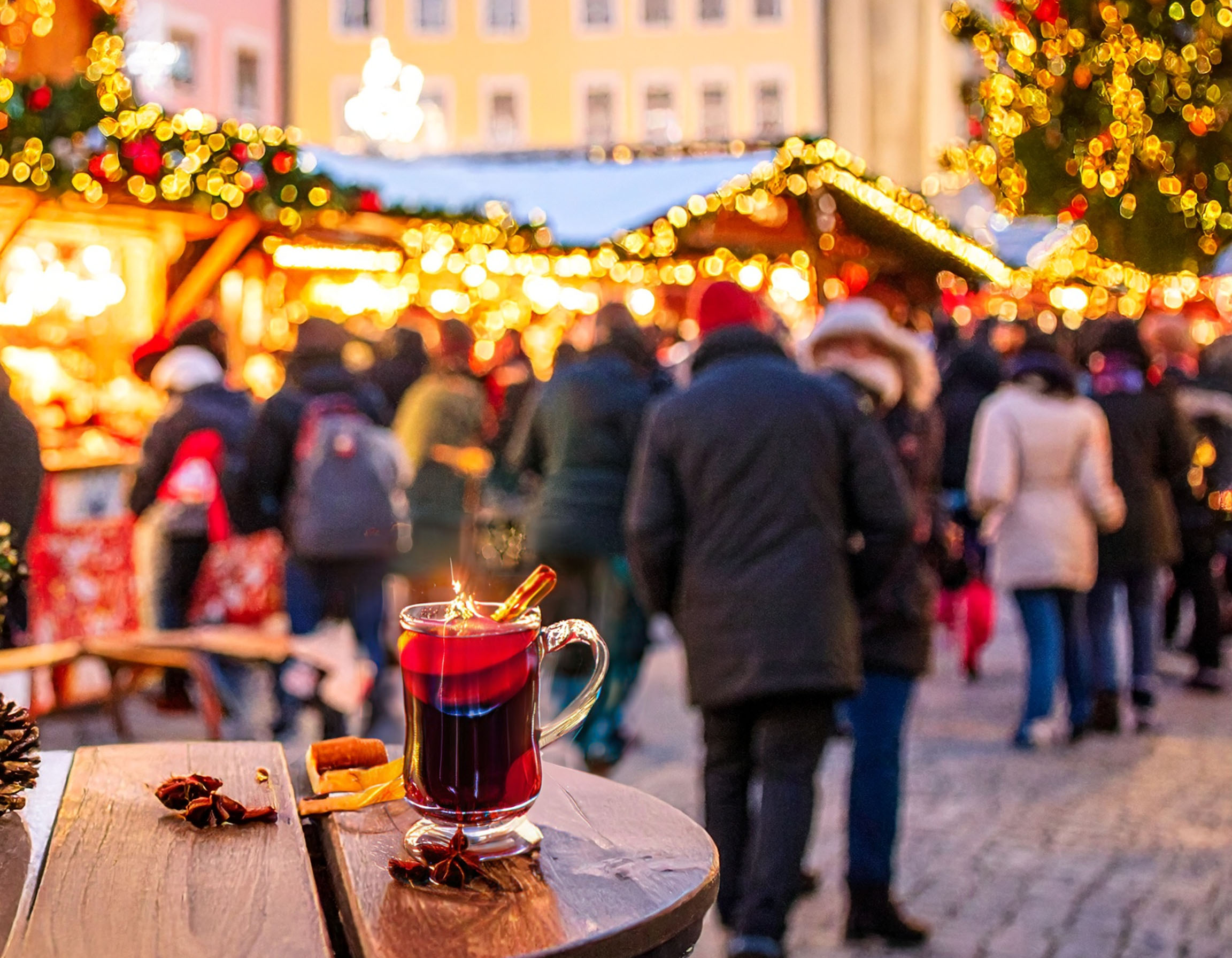 Eine dampfende Tasse Glühwein mit Zimtstange und Sternanis steht im Vordergrund auf einem Holztisch. Im unscharfen Hintergrund erstreckt sich ein belebter Christkindlmarkt bei Abenddämmerung