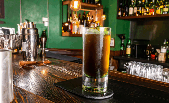 Ein hohes Glas mit einem dunklen Cocktail steht auf einem dunklen Untersetzer auf einer polierten Holztheke. Links davon ist ein silberner Cocktail-Shaker zu sehen