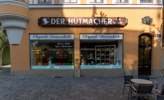 Die Fassade des Hutgeschäfts „Der Hutmacher am Dom", auch bekannt als Hutkönig. Das Gebäude hat eine ockerfarbene Fassade mit zwei großen Schaufenstern, in denen eine Vielzahl eleganter Damen- und Herrenhüte aufgestellt ist. Über den Schaufenstern prangt ein braunes Schild mit dem weißen Schriftzug „Der Hutmacher am Dom" und einem stilisierten Hut