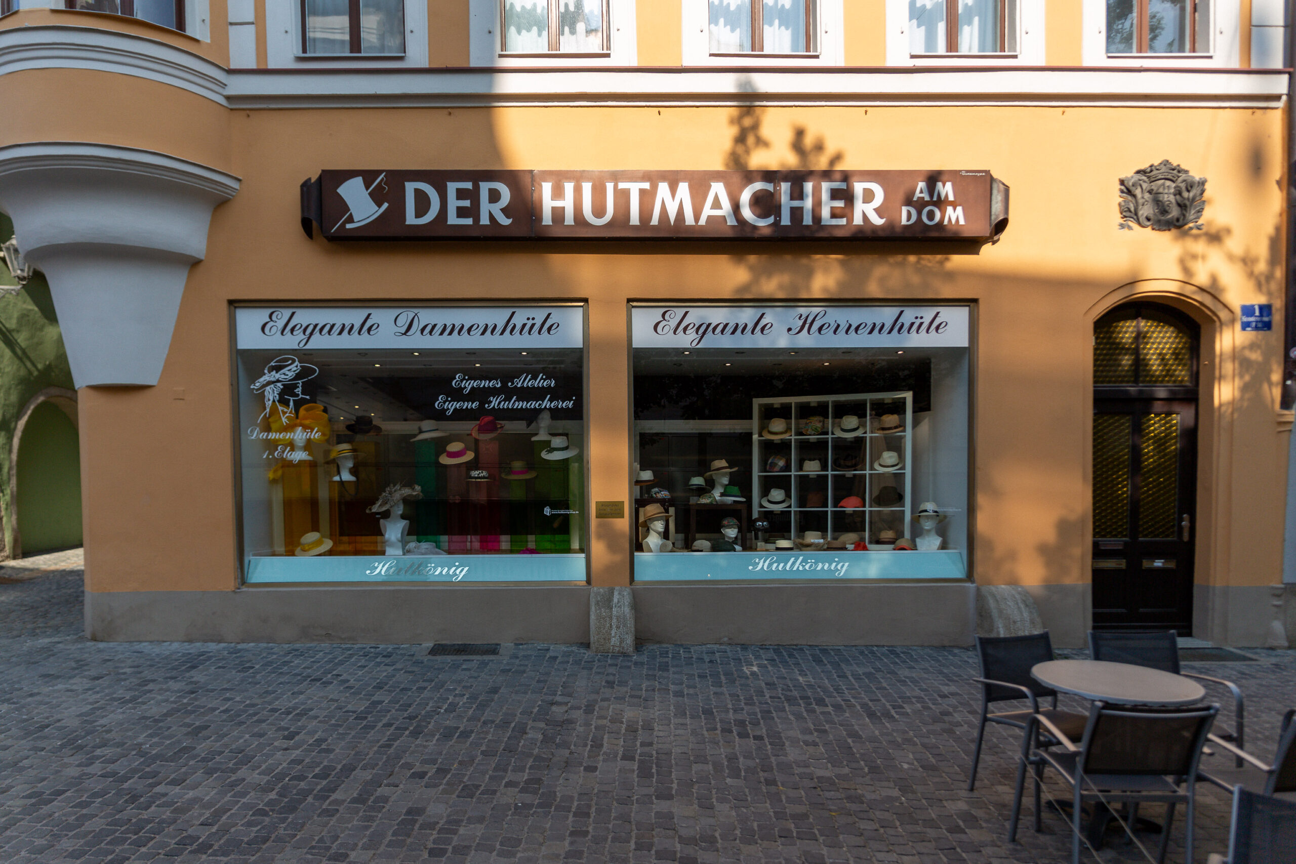 Die Fassade des Hutgeschäfts „Der Hutmacher am Dom", auch bekannt als Hutkönig. Das Gebäude hat eine ockerfarbene Fassade mit zwei großen Schaufenstern, in denen eine Vielzahl eleganter Damen- und Herrenhüte aufgestellt ist. Über den Schaufenstern prangt ein braunes Schild mit dem weißen Schriftzug „Der Hutmacher am Dom" und einem stilisierten Hut