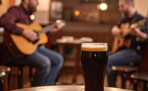 Ein volles Glas dunkles Guinness mit Schaumkrone steht auf einem rustikalen Holztisch im Vordergrund. Im unscharfen Hintergrund sind zwei Männer zu sehen, die auf akustischen Gitarren spielen. Die Beleuchtung ist warm und gedämpft.