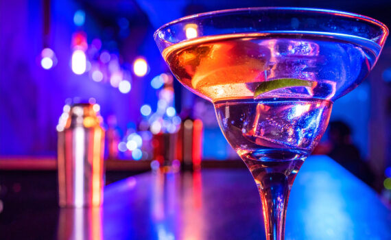 Ein Cocktailglas mit einem klaren Getränk und einer Limettenscheibe steht im Vordergrund auf einer Bar. Die Szene ist in lebhaftes, neonfarbenes Licht getaucht, hauptsächlich in Blau und Rot.