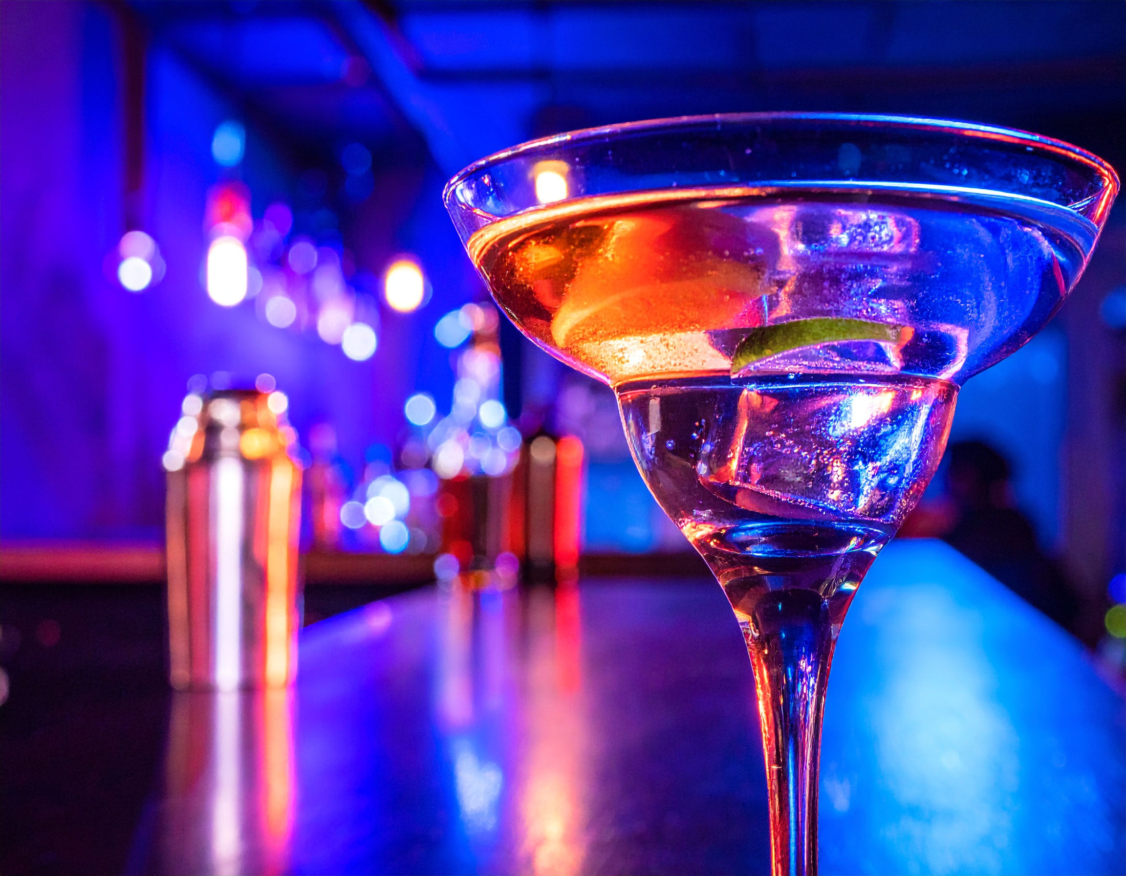 Ein Cocktailglas mit einem klaren Getränk und einer Limettenscheibe steht im Vordergrund auf einer Bar. Die Szene ist in lebhaftes, neonfarbenes Licht getaucht, hauptsächlich in Blau und Rot.