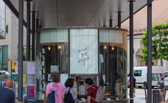 Ein modernes, runder Glaskiosk unter einem Vordach. Auf der Glasfassade ist der Schriftzug „WHAT THE KIOSK?" prominent zu lesen. Im Inneren sind Kaffeeautomaten und Backwaren zu erkennen. Mehrere Personen stehen davor und bestellen.