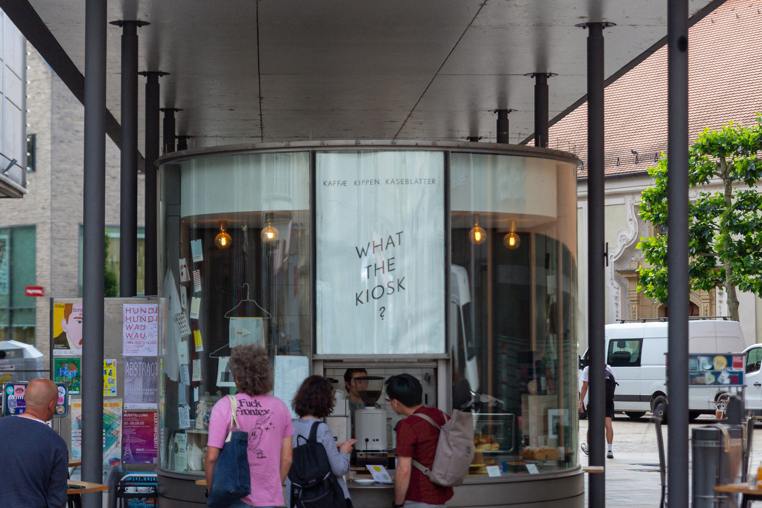 Ein modernes, runder Glaskiosk unter einem Vordach. Auf der Glasfassade ist der Schriftzug „WHAT THE KIOSK?" prominent zu lesen. Im Inneren sind Kaffeeautomaten und Backwaren zu erkennen. Mehrere Personen stehen davor und bestellen.