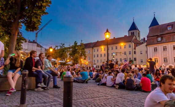Menschen sitzen am am Bismarckplatz in Regensburg am Abend zur Blauen Stunde.