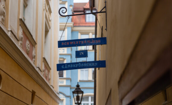 Ein blaues, schmiedeeisernes Hängeschild mit drei horizontalen Tafeln in einer engen Altstadtgasse. Die Tafeln tragen den Schriftzug „schwesternliebe", darunter„in" und ganz unten „Gänsefsschen"