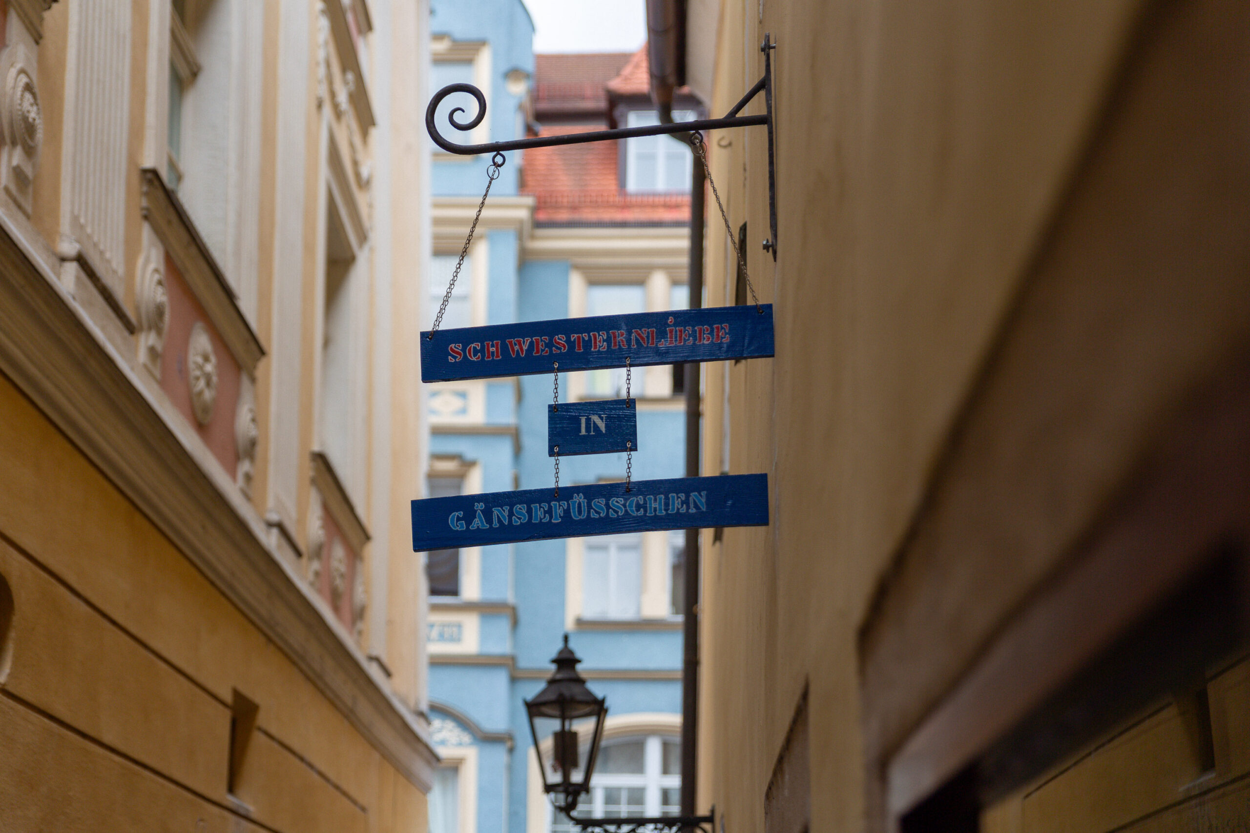 Ein blaues, schmiedeeisernes Hängeschild mit drei horizontalen Tafeln in einer engen Altstadtgasse. Die Tafeln tragen den Schriftzug „schwesternliebe", darunter„in" und ganz unten „Gänsefsschen"
