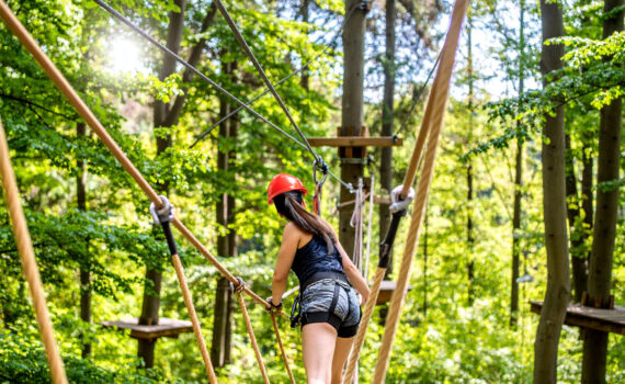 Eine Person mit rotem Schutzhelm und Sicherungsgurt balanciert auf einer Hängebrücke aus Seilen und Holzstegen im Kletterwald.