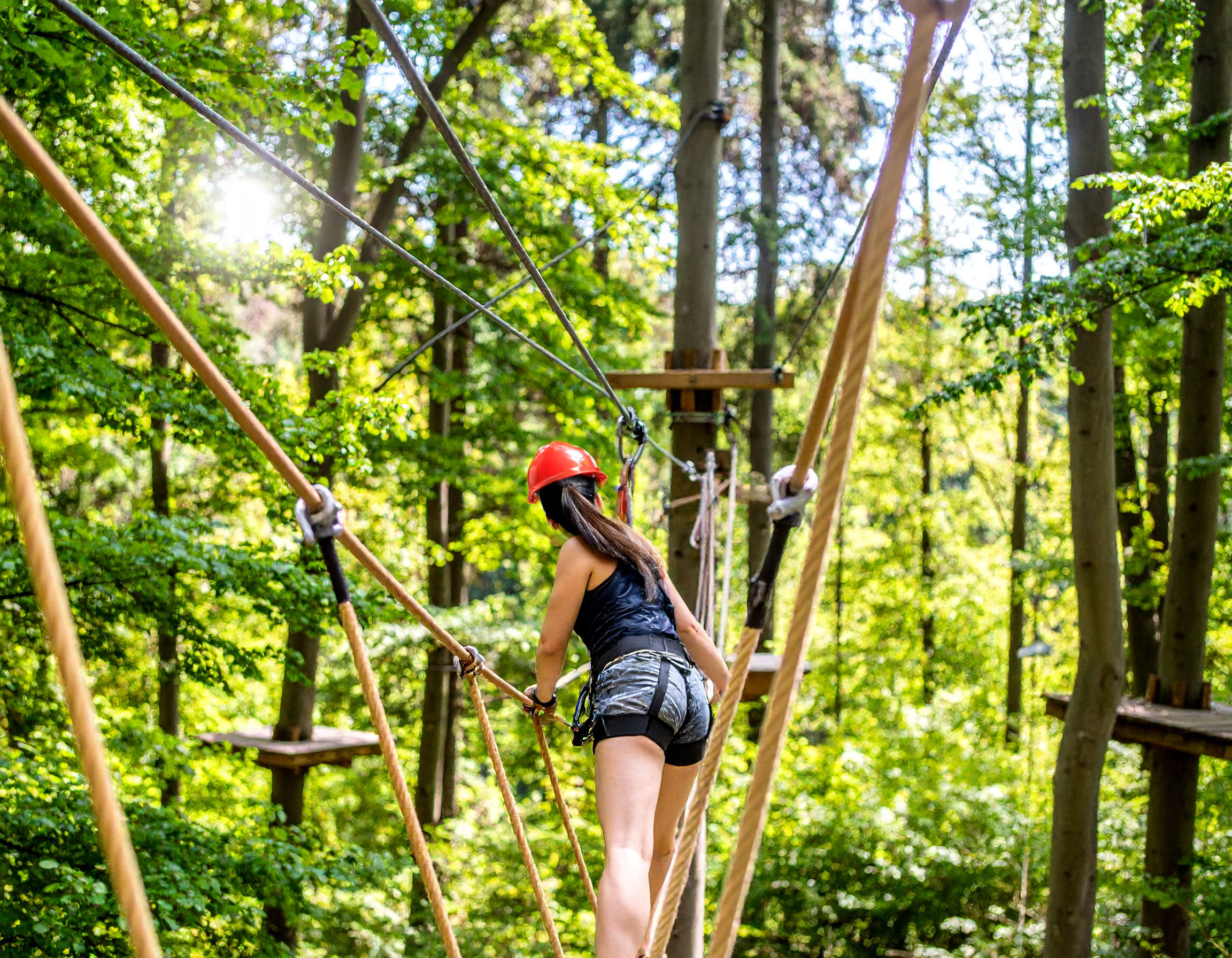 Eine Person mit rotem Schutzhelm und Sicherungsgurt balanciert auf einer Hängebrücke aus Seilen und Holzstegen im Kletterwald.