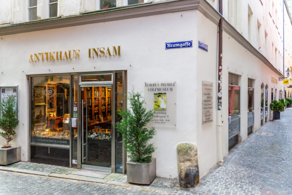 Die Ecke des Antikhauses Insam, das an einer gepflasterten Straße liegt. Das Schaufenster des Geschäfts ist hell beleuchtet und zeigt eine Vielzahl von Antiquitäten wie Schmuck und Silberwaren. Über dem Eingang prangt der goldene Schriftzug „Antikhaus Insam"