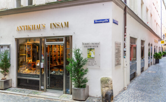 Die Ecke des Antikhauses Insam, das an einer gepflasterten Straße liegt. Das Schaufenster des Geschäfts ist hell beleuchtet und zeigt eine Vielzahl von Antiquitäten wie Schmuck und Silberwaren. Über dem Eingang prangt der goldene Schriftzug „Antikhaus Insam"