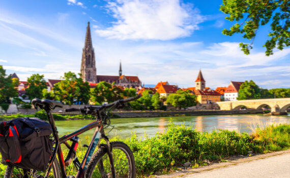 Ein Fahrrad mit Gepäcktasche steht im Vordergrund auf einem Radweg. Im Hintergrund erstreckt sich ein malerischer Blick auf die Stadt Regensburg und die Donau. Der imposante Regensburger Dom mit seinen Türmen und die Steinerne Brücke sind deutlich zu erkennen.