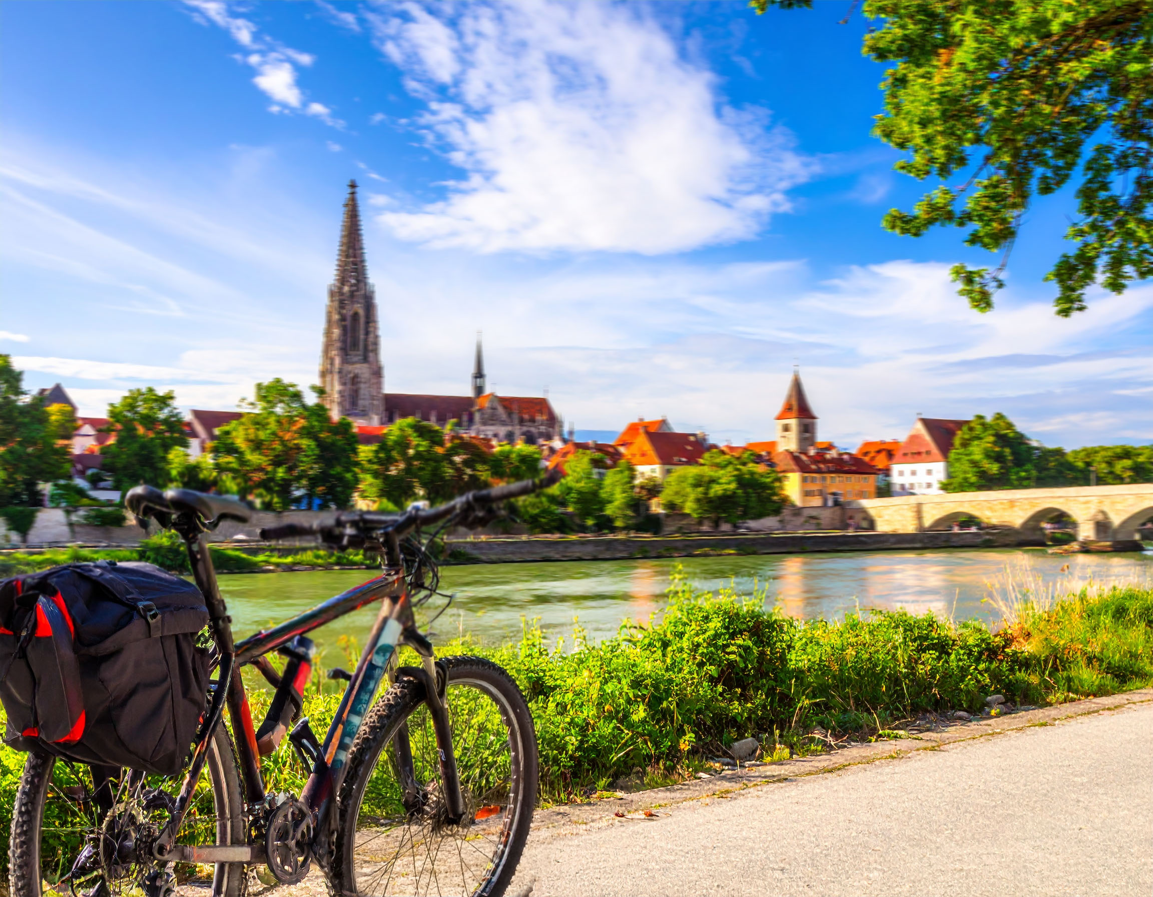 Ein Fahrrad mit Gepäcktasche steht im Vordergrund auf einem Radweg. Im Hintergrund erstreckt sich ein malerischer Blick auf die Stadt Regensburg und die Donau. Der imposante Regensburger Dom mit seinen Türmen und die Steinerne Brücke sind deutlich zu erkennen.